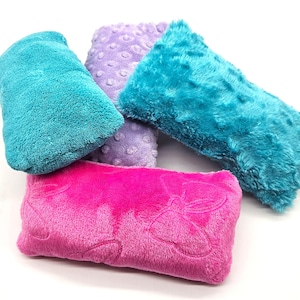 May include: A collection of plush, rectangular pillows in various colors. The pillows are in shades of teal, purple, and pink. The textures vary, with some having a smooth, velvety feel and others a more textured, plush appearance. The pillows are arranged on a white background.