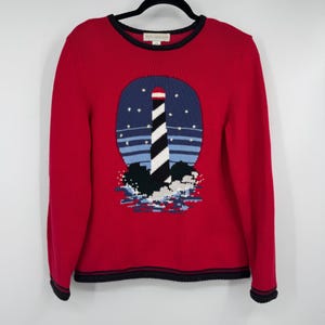 Könnte beinhalten: Roter Pullover mit Leuchtturm-Design. Der Pullover hat einen runden marineblauen Hintergrund mit einem schwarz-weißen Leuchtturm, umgeben von Sternen und Wellen. Der Pullover hat eine schwarze Bordüre um den Halsausschnitt, die Manschetten und den Saum.