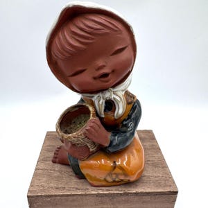 May include: A ceramic figurine of a girl wearing a white bonnet and a yellow dress, holding a brown basket. The figurine is sitting on a wooden base.