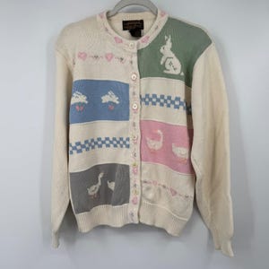 May include: Cream-colored cardigan sweater with a button-up front. The sweater features a patchwork design with pastel blue, pink, green, and gray blocks. Each block has a different design, including bunnies, geese, and checkered patterns. The sweater has long sleeves and a round neckline.