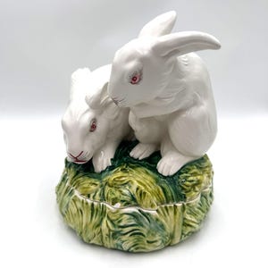 May include: A white ceramic figurine of two rabbits sitting on a green base. The rabbits have red eyes and are in a playful pose.