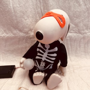 May include: A Snoopy figurine dressed in a black skeleton costume with white bone detailing. The figurine wears an orange eye mask and holds a glowing white ball. A black battery pack is connected to the ball.