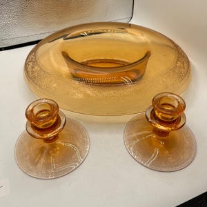 May include: A set of three amber glass pieces, including a large, oval-shaped bowl with a smaller bowl inside and two candle holders with a decorative etched pattern.