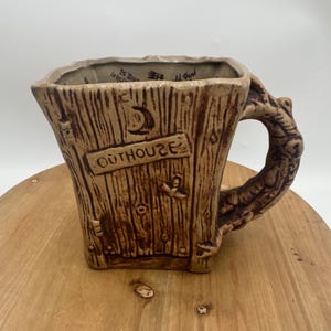 May include: A brown ceramic mug with a square shape and a handle designed to look like a tree branch. The mug has a textured surface resembling wood grain and features the word "OUTHOUSE" on the front.