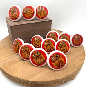 May include: A collection of white ceramic cabinet knobs with a red border and a detailed owl design in orange and black. The knobs are displayed on a wooden surface, showcasing their decorative appeal.