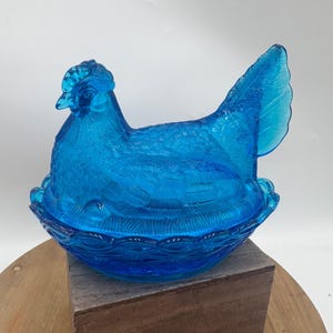 May include: A blue glass hen-shaped dish with a detailed textured design. The hen is sitting on a nest-shaped base. The glass is a vibrant shade of blue, and the design includes feather and basket weave details.