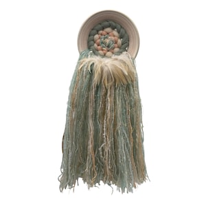 May include: A woven wall hanging with a circular design in shades of green, pink, and white. The hanging has long, flowing strands of yarn in shades of green and beige.