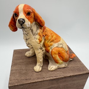 Może przedstawiać: Ceramiczna figurka psa w pozycji siedzącej. Pies ma biało-pomarańczowe umaszczenie z białym paskiem na twarzy. Figurka stoi na drewnianym klocku.