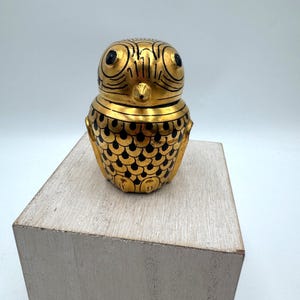 May include: A gold-colored owl-shaped decorative container with black detailing. The owl has large, round eyes and a beak. The body is adorned with a scale-like pattern in black. The container is sitting on a light-colored wooden block.