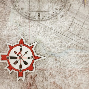 May include: A compass patch with a red and white design, featuring arrows and directional indicators, rests on a detailed topographic map. A clear plastic protractor and a map scale are also visible, suggesting navigation or exploration.