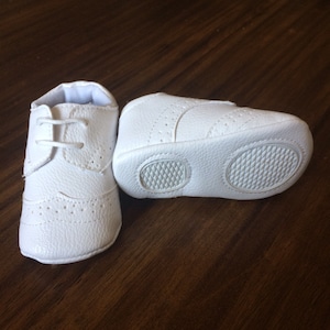 K&ouml;nnte beinhalten: Ein Paar wei&szlig;e Lederschuhe f&uuml;r Babys mit Schn&uuml;rsenkeln. Die Schuhe haben ein strukturiertes Design und eine Gummisohle.