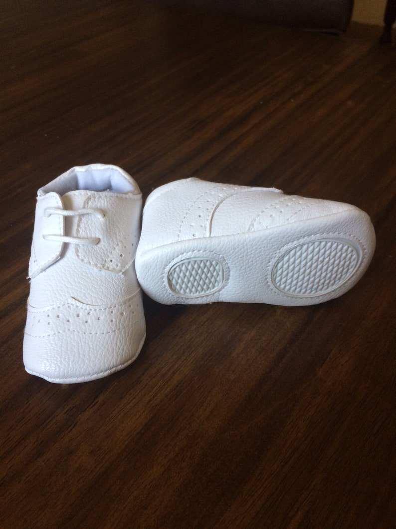 K&ouml;nnte beinhalten: Ein Paar wei&szlig;e Lederschuhe f&uuml;r Babys mit Schn&uuml;rsenkeln. Die Schuhe haben ein strukturiertes Design und eine Gummisohle.