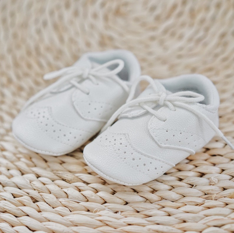 K&ouml;nnte beinhalten: Ein Paar wei&szlig;e Lederschuhe f&uuml;r Babys mit Schn&uuml;rsenkeln. Die Schuhe haben ein perforiertes Design und befinden sich auf einer gewebten Oberfl&auml;che.