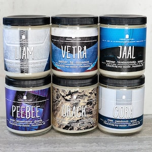 May include: Six glass candle jars with black lids and labels. The labels have different names and scents. The candles are arranged in two rows of three. The candles are on a light wood surface.