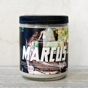 May include: A glass jar candle with a black lid. The label features the name "MARCUS" in large white letters, with additional text indicating it's a cream leather wrapped steel topper soy candle, 7.5oz/213g. The label has a photo of a person.