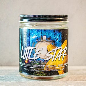 May include: A glass candle jar with a black lid. The label features a blue and yellow design with the text "EMBERS AND ASHES COMPANY" and "LITTLE STAR". The text below reads "small forest. currents of time. quiet secrets. 7.5oz/213g soy candle. Pendleton, IN."