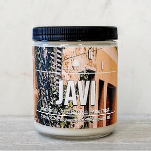 May include: A glass jar candle with a black lid. The label features the name "JAVI" in large white letters, with "sangria, cinnamon, sea mist" below. The label also includes a scenic image of a building.