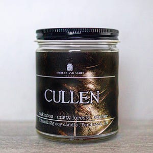 May include: A glass candle jar with a black lid and a label that reads "CULLEN" in white lettering. The label also includes the text "EMBERS AND ASHES COMPANY" and "oakmoss, misty forests, amber 7.5oz/213g soy candle. Pendleton, IN".