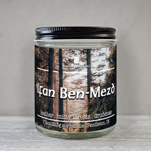 May include: A glass jar candle with a black lid. The label features the text "Fan Ben-Mezd" over a forest scene. The label also includes the text "leather, misty forests, drudenae" and "7.5oz/213g soy candle".