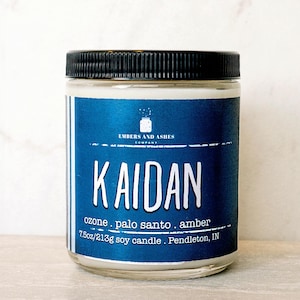 May include: A blue and white soy candle jar with a black lid. The label reads "Embers and Ashes Company" and "Kaidan" in a white font. The scent description is "ozone, palo santo, amber". The candle is 7.5 oz/213g and made in Pendleton, IN.