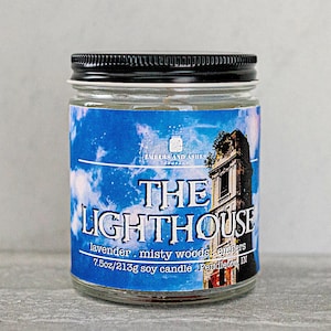 May include: A glass candle jar with a black lid. The label is blue with white clouds and a building with a window. The text on the label reads "THE LIGHTHOUSE lavender. misty woods. embers 7.5oz/213g soy candle Pendleton IN".