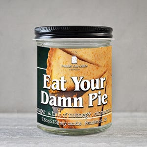 May include: A clear glass jar candle with a black lid. The label reads "Eat Your Damn Pie" with text about ingredients. The candle is 7.5oz (213g) and made of soy wax. The label features a pie graphic.