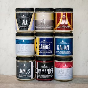 May include: Nine glass jar candles with black lids and labels featuring names like "TALI," "EDI," and "JAVIK." Each label has a unique color scheme and scent description. The candles are arranged in three rows.