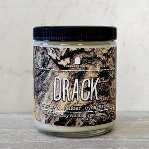 May include: A glass jar candle with a black lid and a label that says "Drack" in white lettering. The label also says "oakmoss, amber, cybernetics" and "7 oz/213g soy candle, Pendleton, IN". The jar has a brown and white camouflage pattern.