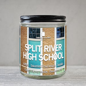 May include: A glass jar candle with a black lid. The label features the text "SPLIT RIVER HIGH SCHOOL" in white, with the words "coffee, haunted hallways, books" below. The label has a brick and window design.