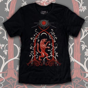 May include: Black t-shirt with a red wolf design. The wolf is standing on its hind legs inside a circular frame with a sun and moon above it. The design is surrounded by vines and leaves.