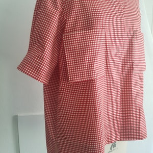 Könnte beinhalten: Kurzarmhemd aus rotem und weißem Gingham mit einer Tasche auf der Vorderseite. Das Hemd hat einen leicht abgerundeten Ausschnitt und einen geraden Saum. Das Etikett auf dem Hemd lautet "Mori Plan Made in Thailand".