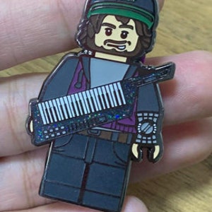 May include: A black and silver enamel pin of a Lego minifigure wearing a black and green hat with the letters "MFG" on it. The minifigure is holding a silver and black keyboard.