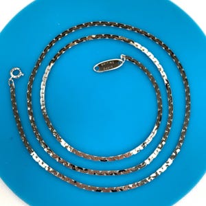 May include: A silver-toned metal chain necklace featuring a flat, interlocking link design. The necklace includes a small, oval-shaped metal tag engraved with the text Whiting Davis and a circular spring ring clasp at the end of the chain.