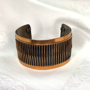 May include: A copper cuff bracelet with a geometric pattern. The bracelet is made of a dark copper metal with a black enamel design.