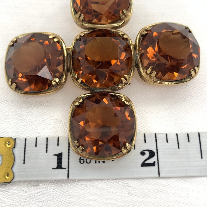 May include: A gold-toned brooch with five brown, faceted glass stones arranged in a cross shape. The brooch measures approximately 1 1/2 inches in length.