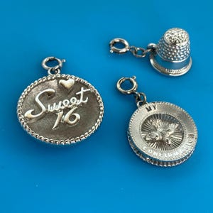 May include: Three silver-toned metal charms including a round charm with the text Sweet 16 and a small heart, a round charm featuring a dove and the text My Confirmation, and a small thimble charm, each attached to a circular jump ring connector.