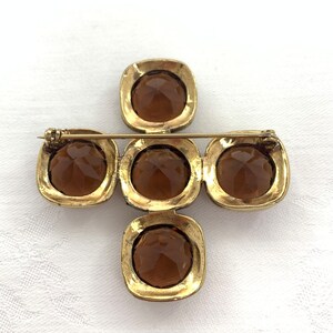 May include: A gold-toned brooch in the shape of a cross with five amber-colored glass stones. The brooch has a pin on the back for attaching to clothing.