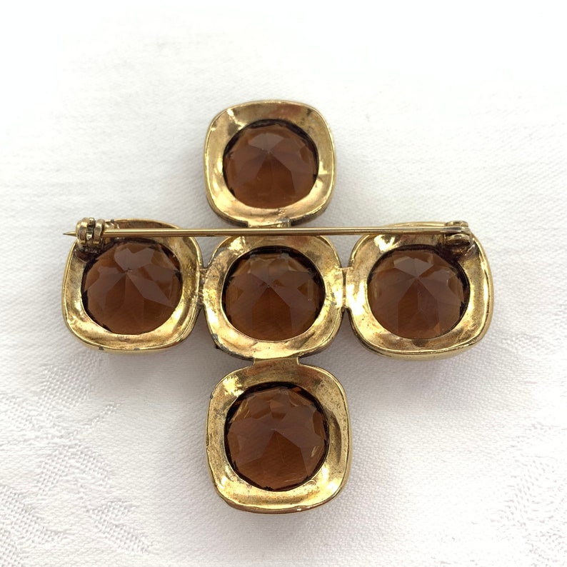 May include: A gold-toned brooch in the shape of a cross with five amber-colored glass stones. The brooch has a pin on the back for attaching to clothing.