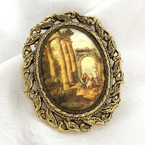 May include: A gold-toned oval brooch with a detailed leaf design. The center features a cameo with a painted scene of an ancient Roman ruin.