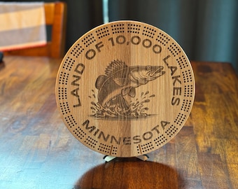May include: A circular wooden cribbage board featuring an engraved leaping fish design. The text "LAND OF 10,000 LAKES" and "MINNESOTA" are etched around the fish. The board is designed with numerous holes for pegs.