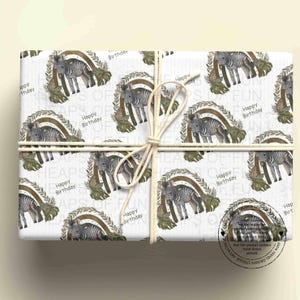 May include: White wrapping paper featuring a repeating pattern of zebras standing under brown rainbows with green leaves. The text "Happy Birthday" is printed near each zebra. A thick white string is tied around the centre of the paper.