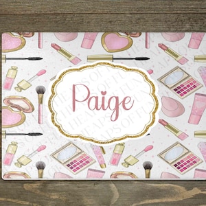 May include: A rectangular glass cutting board featuring a repeating pattern of pink and gold makeup items including lipstick, mascara, and eyeshadow palettes. A central white scalloped label with a gold glitter border displays the name Paige.