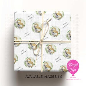 May include: A square gift box wrapped in white paper featuring a repeating pattern of green leaves, gold number eights, and small zebras. The text Happy Birthday is printed in black. The box is tied with a light beige string in a cross pattern.