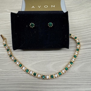 May include: A gold-tone tennis bracelet and a pair of matching stud earrings featuring alternating round-cut emerald green and clear white stones. The jewellery set is presented in a black display box with the brand name AVON printed in gold lettering.