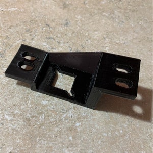May include: A black, 3D-printed bracket with a rectangular body and two angled side extensions. Each extension has two circular holes. The center features a square cutout with a unique shape. The bracket is designed for mounting or support.