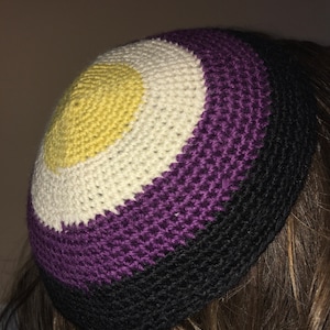Könnte beinhalten: Eine gehäkelte Kippa mit einem Farbmuster in Gelb, Weiß, Lila und Schwarz. Die Kippa ist eine traditionelle jüdische Kopfbedeckung.
