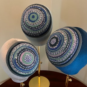 Può includere: Tre kippa all'uncinetto con motivi circolari blu, verdi e rosa. Le kippa sono esposte su supporti dorati.
