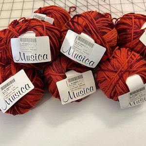 May include: Six balls of red and burgundy yarn with a metallic sheen. Each ball has a white label with the text "L-M-0061-1 Musica Price 9.95 Capital Yarns 703 263-9744 Musica".