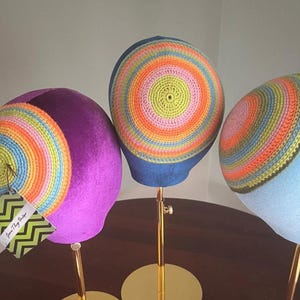 Op de afbeelding: Drie kleurrijke gehaakte hoeden. De hoeden hebben concentrische cirkels in levendige kleuren, waaronder geel, roze, oranje en blauw. Elke hoed is gemonteerd op een gouden standaard met een ronde basis. Eén hoed heeft een label met "Jane Thy Vintage".