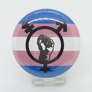May include: A round button with a transgender pride flag design. The flag is blue, pink, white, and light blue. A black fist is inside a circle with a transgender symbol.